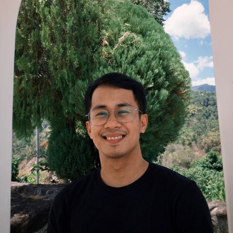 Rj smiling, in glasses and a black t-shirt, in front of a cypress tree